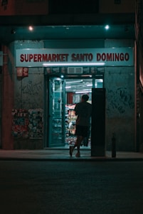 A person is entering a small, illuminated supermarket at night. The store, named Supermarket Santo Domingo, has a neon glow with the sign displayed above. Graffiti and posters cover the walls around the entrance, creating an urban atmosphere.