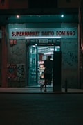 A person is entering a small, illuminated supermarket at night. The store, named Supermarket Santo Domingo, has a neon glow with the sign displayed above. Graffiti and posters cover the walls around the entrance, creating an urban atmosphere.