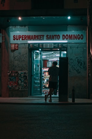 A person is entering a small, illuminated supermarket at night. The store, named Supermarket Santo Domingo, has a neon glow with the sign displayed above. Graffiti and posters cover the walls around the entrance, creating an urban atmosphere.