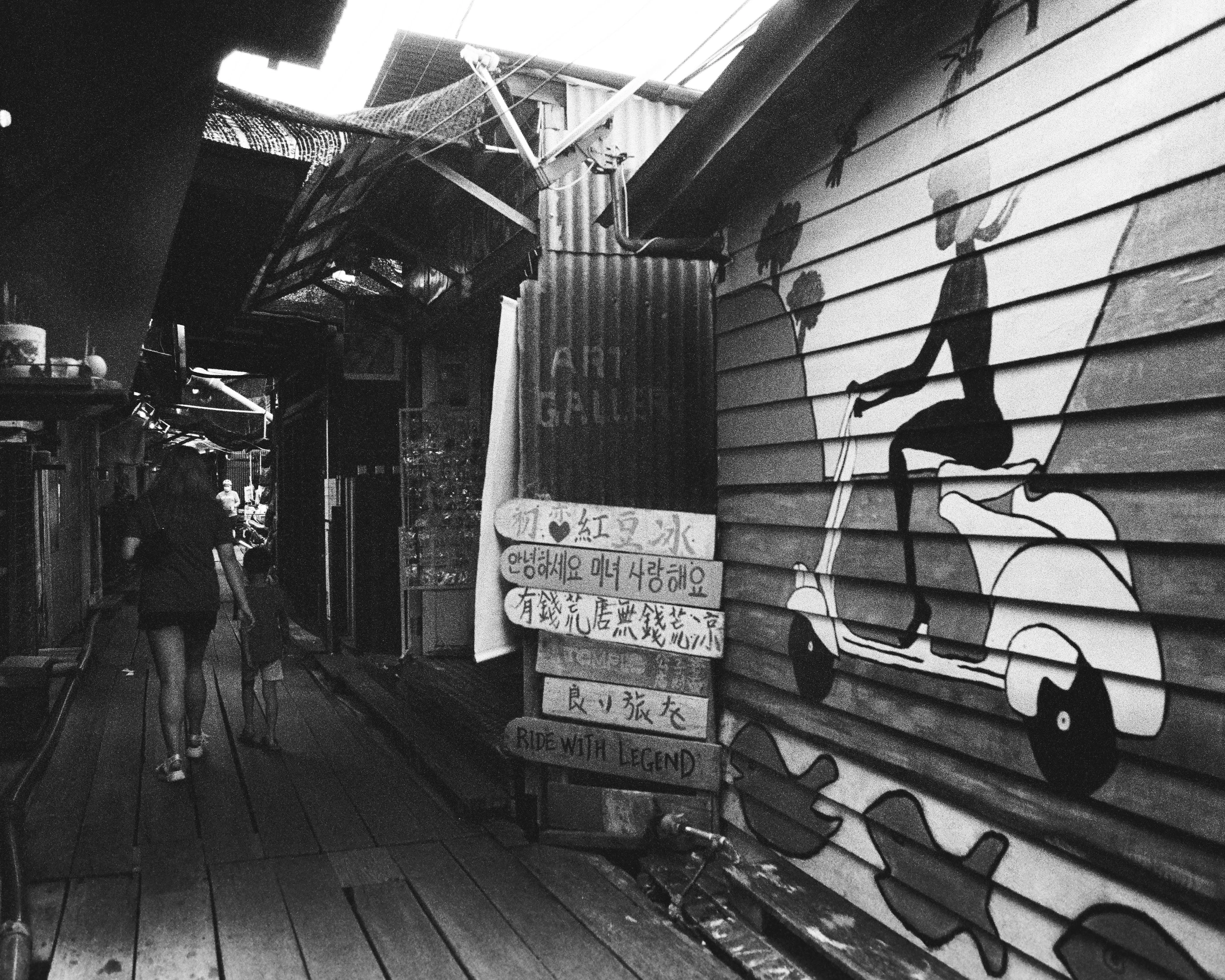Black-and-white view down a wooden stilt-house jetty alley with a painted scooter mural and hand-lettered signs.
