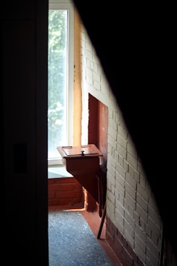 A narrow corridor features an old red metal mailbox mounted on a brick wall adjacent to a bright window. Light from the window illuminates parts of the scene, casting soft shadows on the floor and walls. The bricks are painted a combination of beige and red-brown, accentuating the rustic atmosphere of the space.