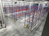 An industrial warehouse with multiple rows of blue and red metal storage racks. The floor is concrete, and the warehouse is well-lit with high ceiling lights. A fire extinguisher is located at the lower-left corner of the warehouse.