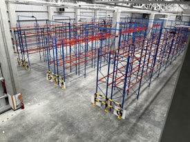 An industrial warehouse with multiple rows of blue and red metal storage racks. The floor is concrete, and the warehouse is well-lit with high ceiling lights. A fire extinguisher is located at the lower-left corner of the warehouse.