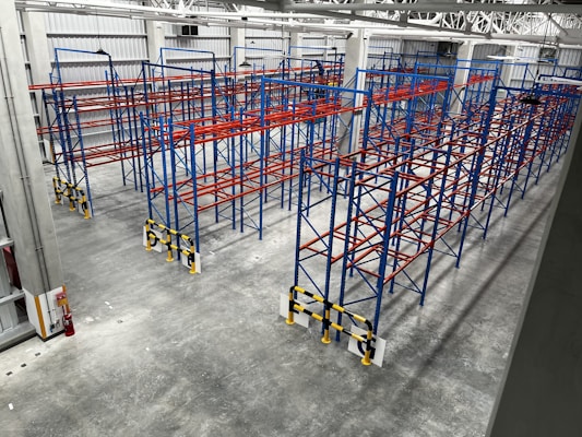 An industrial warehouse with multiple rows of blue and red metal storage racks. The floor is concrete, and the warehouse is well-lit with high ceiling lights. A fire extinguisher is located at the lower-left corner of the warehouse.