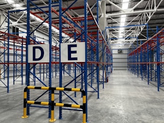 A large, empty warehouse with metal shelving units arranged in parallel rows. The shelves are constructed with blue and red beams. The floor is made of smooth, gray concrete, and overhead lights illuminate the space. Signs with letters 'D' and 'E' are prominently displayed on the racks near the foreground.