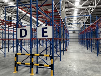 A large, empty warehouse with metal shelving units arranged in parallel rows. The shelves are constructed with blue and red beams. The floor is made of smooth, gray concrete, and overhead lights illuminate the space. Signs with letters 'D' and 'E' are prominently displayed on the racks near the foreground.