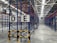 A large, empty warehouse with metal shelving units arranged in parallel rows. The shelves are constructed with blue and red beams. The floor is made of smooth, gray concrete, and overhead lights illuminate the space. Signs with letters 'D' and 'E' are prominently displayed on the racks near the foreground.