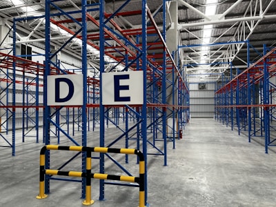 A large, empty warehouse with metal shelving units arranged in parallel rows. The shelves are constructed with blue and red beams. The floor is made of smooth, gray concrete, and overhead lights illuminate the space. Signs with letters 'D' and 'E' are prominently displayed on the racks near the foreground.