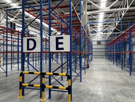 A large, empty warehouse with metal shelving units arranged in parallel rows. The shelves are constructed with blue and red beams. The floor is made of smooth, gray concrete, and overhead lights illuminate the space. Signs with letters 'D' and 'E' are prominently displayed on the racks near the foreground.