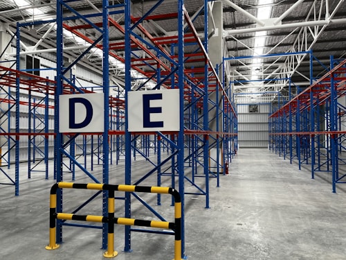 A large, empty warehouse with metal shelving units arranged in parallel rows. The shelves are constructed with blue and red beams. The floor is made of smooth, gray concrete, and overhead lights illuminate the space. Signs with letters 'D' and 'E' are prominently displayed on the racks near the foreground.