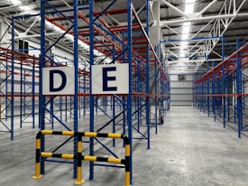 A large, empty warehouse with metal shelving units arranged in parallel rows. The shelves are constructed with blue and red beams. The floor is made of smooth, gray concrete, and overhead lights illuminate the space. Signs with letters 'D' and 'E' are prominently displayed on the racks near the foreground.