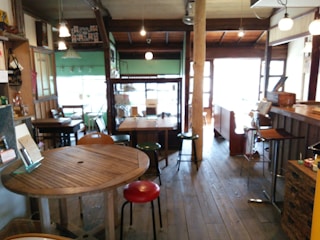 A cozy café interior featuring wooden tables and chairs with a rustic, homey atmosphere. The lighting is soft with several hanging lamps, and the floor is made of wooden planks. The space appears intimate and inviting, combining elements of vintage and contemporary design.