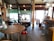 A cozy café interior featuring wooden tables and chairs with a rustic, homey atmosphere. The lighting is soft with several hanging lamps, and the floor is made of wooden planks. The space appears intimate and inviting, combining elements of vintage and contemporary design.