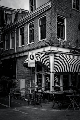 A street scene featuring an outdoor seating area of a café with striped awnings. The building is made of brick and has large windows with various signs on the exterior. There are tables and chairs placed outside, and a street sign is visible in the foreground.