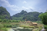 A serene rural landscape features lush green mountains under a partially cloudy sky. A quaint village with thatched-roof houses sits in the foreground, surrounded by vegetation. A tranquil body of water reflects the greenery and sky, with patches of water lilies adding to the tranquility.