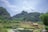 A serene rural landscape features lush green mountains under a partially cloudy sky. A quaint village with thatched-roof houses sits in the foreground, surrounded by vegetation. A tranquil body of water reflects the greenery and sky, with patches of water lilies adding to the tranquility.