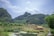 A serene rural landscape features lush green mountains under a partially cloudy sky. A quaint village with thatched-roof houses sits in the foreground, surrounded by vegetation. A tranquil body of water reflects the greenery and sky, with patches of water lilies adding to the tranquility.