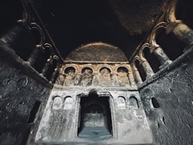 A dimly lit ancient stone structure features arches and niches on its walls, creating a sense of historical depth and mystery. The surfaces show signs of wear and age, adding to the ancient atmosphere.