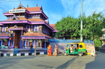 A traditional Indian temple with vibrant purple and orange hues and intricate architectural details. People dressed in traditional clothing are gathered around, with some sitting on a low wall in front of the temple. A green and yellow auto-rickshaw is parked nearby under a clear blue sky, with lush green foliage and decorative elements visible.