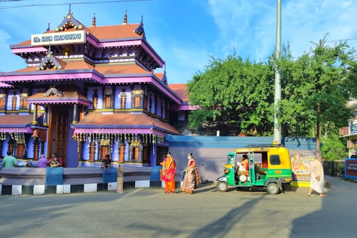 A traditional Indian temple with vibrant purple and orange hues and intricate architectural details. People dressed in traditional clothing are gathered around, with some sitting on a low wall in front of the temple. A green and yellow auto-rickshaw is parked nearby under a clear blue sky, with lush green foliage and decorative elements visible.