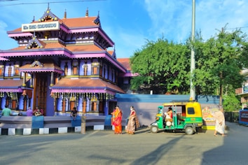 A traditional Indian temple with vibrant purple and orange hues and intricate architectural details. People dressed in traditional clothing are gathered around, with some sitting on a low wall in front of the temple. A green and yellow auto-rickshaw is parked nearby under a clear blue sky, with lush green foliage and decorative elements visible.