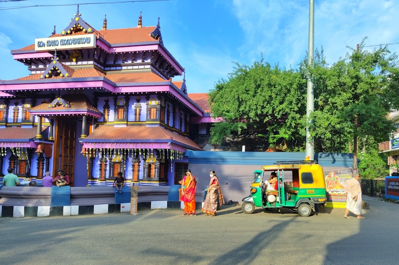 A traditional Indian temple with vibrant purple and orange hues and intricate architectural details. People dressed in traditional clothing are gathered around, with some sitting on a low wall in front of the temple. A green and yellow auto-rickshaw is parked nearby under a clear blue sky, with lush green foliage and decorative elements visible.