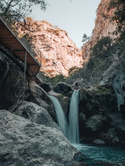 A serene natural scene featuring a small waterfall cascading into a clear pool surrounded by large rocks. The waterfall is framed by rugged cliffs with sparse vegetation. A pathway bridge runs along the cliff edge in the upper left corner of the image, leading the eye along the landscape. The atmosphere is calm and peaceful, with an overall earthy and rugged aesthetic.