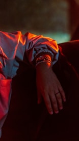 A hand resting on a dark surface, illuminated by a warm, reddish light. The wrist is adorned with a shiny watch and a bracelet. The sleeve of a light-colored jacket is slightly rolled up, and the focus is mainly on the hand and wrist area.
