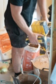 A person wearing a black shirt and shorts is using a yellow power drill mixer to blend a substance in a large container. Surrounding the person are bags of building materials and various construction tools on a floor covered with protective material.