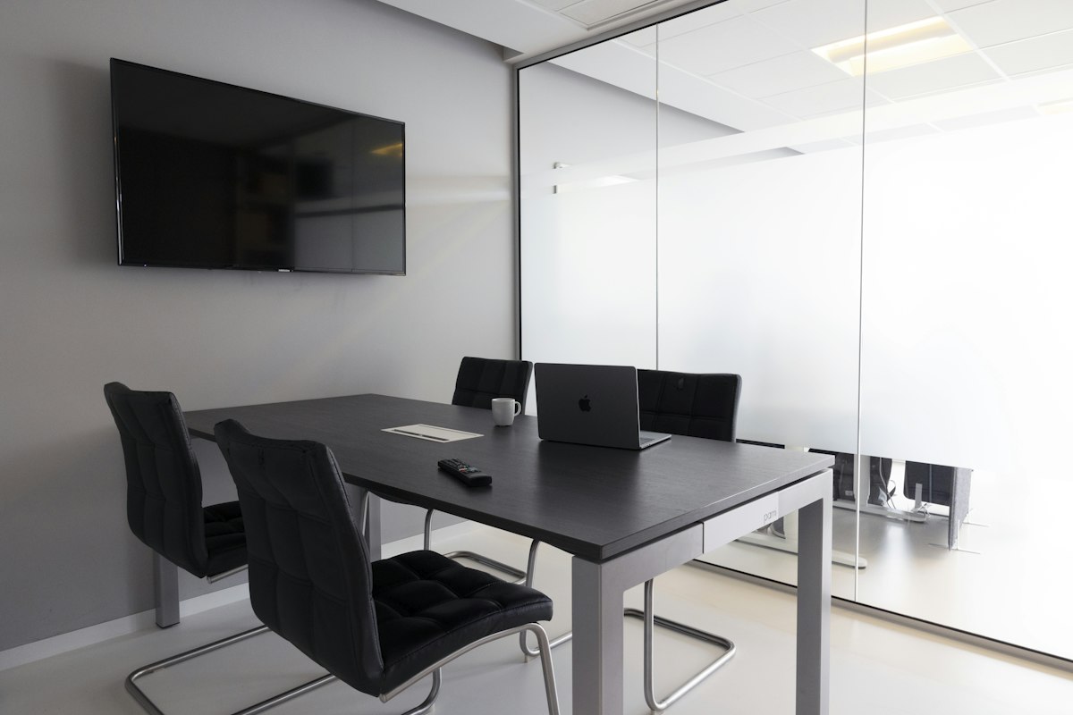 Petite salle de réunion avec une table noire, quatre chaises, un ordinateur portable ouvert et un écran fixé au mur, séparée du couloir par une paroi vitrée