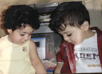Two young children are sitting side by side, looking down. The child on the left has curly hair and is wearing a light yellow sleeveless top. The child on the right has short hair and is wearing a red checkered shirt over a white T-shirt. Behind them, there is a computer monitor with various items stored above it.