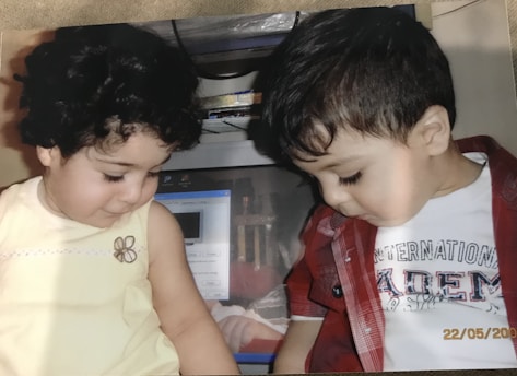 Two young children are sitting side by side, looking down. The child on the left has curly hair and is wearing a light yellow sleeveless top. The child on the right has short hair and is wearing a red checkered shirt over a white T-shirt. Behind them, there is a computer monitor with various items stored above it.