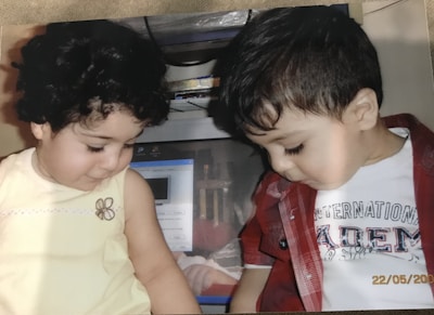 Two young children are sitting side by side, looking down. The child on the left has curly hair and is wearing a light yellow sleeveless top. The child on the right has short hair and is wearing a red checkered shirt over a white T-shirt. Behind them, there is a computer monitor with various items stored above it.