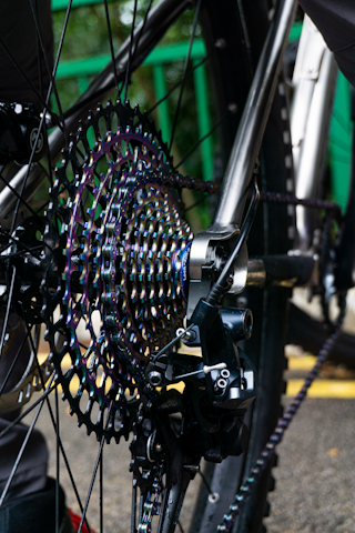 Close-up of a bicycle's rear cassette and derailleur, showing the intricately designed gears with a metallic, iridescent finish. The metal frame and spokes are visible, along with the bike chain. The background includes blurred green railing and a hint of pavement.