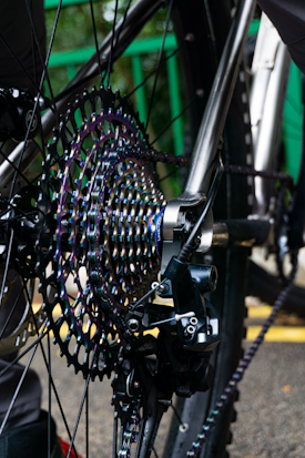 Close-up of a bicycle's rear cassette and derailleur, showing the intricately designed gears with a metallic, iridescent finish. The metal frame and spokes are visible, along with the bike chain. The background includes blurred green railing and a hint of pavement.