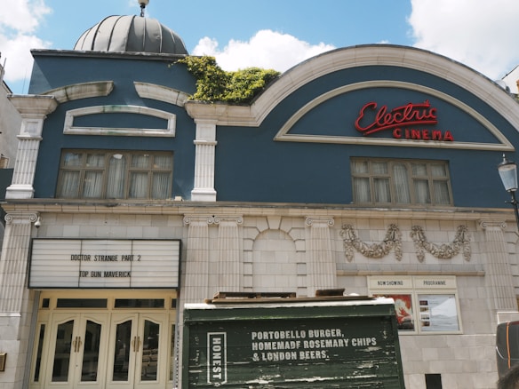 An elegant cinema building with a dome and decorative facade features a vintage red neon sign reading 'Electric Cinema.' The marquee lists the films 'Doctor Strange Part 2' and 'Top Gun Maverick.' A poster display is visible near the entrance, and a green kiosk in the foreground advertises burgers, chips, and beers.