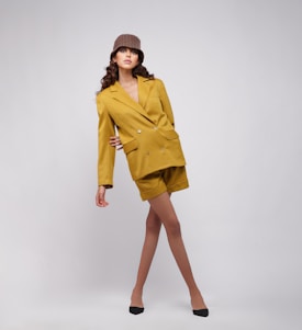 A fashionable model poses confidently wearing a mustard yellow blazer and shorts. She is accessorized with a knitted brown hat and black shoes, set against a minimalist gray background.