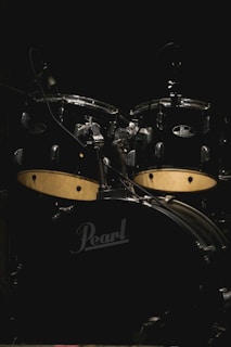 A drum set prominently displayed against a dark background, featuring a black finish with prominent logos. The kit includes several drums and cymbals, well-lit to highlight details.