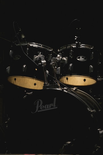 A drum set prominently displayed against a dark background, featuring a black finish with prominent logos. The kit includes several drums and cymbals, well-lit to highlight details.