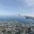 An aerial view of a coastline with a clear blue sea, sprawling agricultural fields, and clusters of buildings. The wing of an airplane is visible, indicating the photo was taken from inside the aircraft.