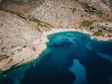 An aerial view captures a secluded beach nestled between rugged rocky cliffs and crystal-clear turquoise waters. Sun umbrellas line the sandy shore, indicating a small, organized area for visitors. The landscape is characterized by dry, rocky terrain with sparse vegetation extending up the hillside.