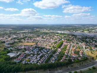 Aerial view of a suburban area with rows of houses and green spaces interspersed throughout. The landscape extends towards the horizon, with patches of forest and a large parking lot visible in the background. The sky is partly cloudy, with fluffy white clouds scattered across a blue sky.