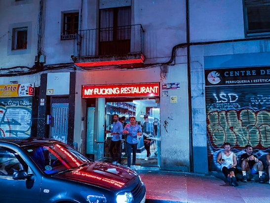 A restaurant entrance with a bright red neon sign displaying bold letters, surrounded by urban graffiti-covered walls. Several people stand near the entrance, with a parked car reflecting the neon light in the foreground. The setting appears to be in a lively urban area during the evening.