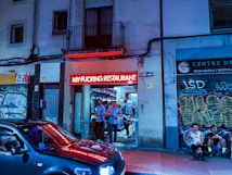 A restaurant entrance with a bright red neon sign displaying bold letters, surrounded by urban graffiti-covered walls. Several people stand near the entrance, with a parked car reflecting the neon light in the foreground. The setting appears to be in a lively urban area during the evening.