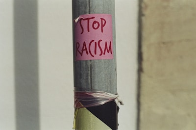 A metal pole with a pink sign attached to it, displaying the handwritten message 'Stop Racism'. The pole appears to be located outdoors, with a blurred background showing a wall.