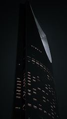 A tall, modern skyscraper with an angular, sleek design is illuminated against a dark background. The building's facade is dotted with lit windows, creating a pattern of warm light against the cool tones of the structure. The architecture displays an elegant curve and sharp edges.
