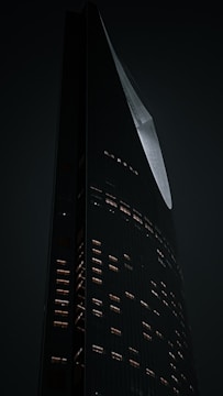 A tall, modern skyscraper with an angular, sleek design is illuminated against a dark background. The building's facade is dotted with lit windows, creating a pattern of warm light against the cool tones of the structure. The architecture displays an elegant curve and sharp edges.