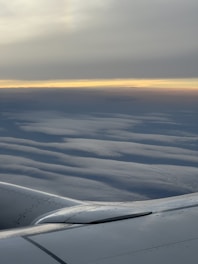 An airplane wing with a smooth, metallic surface is visible against a backdrop of layered, wavy clouds stretching to the horizon. A subtle gradient of colors from light blue to warm orange hints at either sunrise or sunset, creating a serene and calm atmosphere.