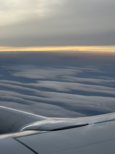 An airplane wing with a smooth, metallic surface is visible against a backdrop of layered, wavy clouds stretching to the horizon. A subtle gradient of colors from light blue to warm orange hints at either sunrise or sunset, creating a serene and calm atmosphere.
