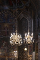 Elaborate chandeliers hang from the ceiling in an ornate setting filled with intricate religious artwork and patterns. The backdrop includes vibrant paintings depicting religious figures and symbols. The atmosphere is richly decorated, giving a sense of historical and cultural significance.